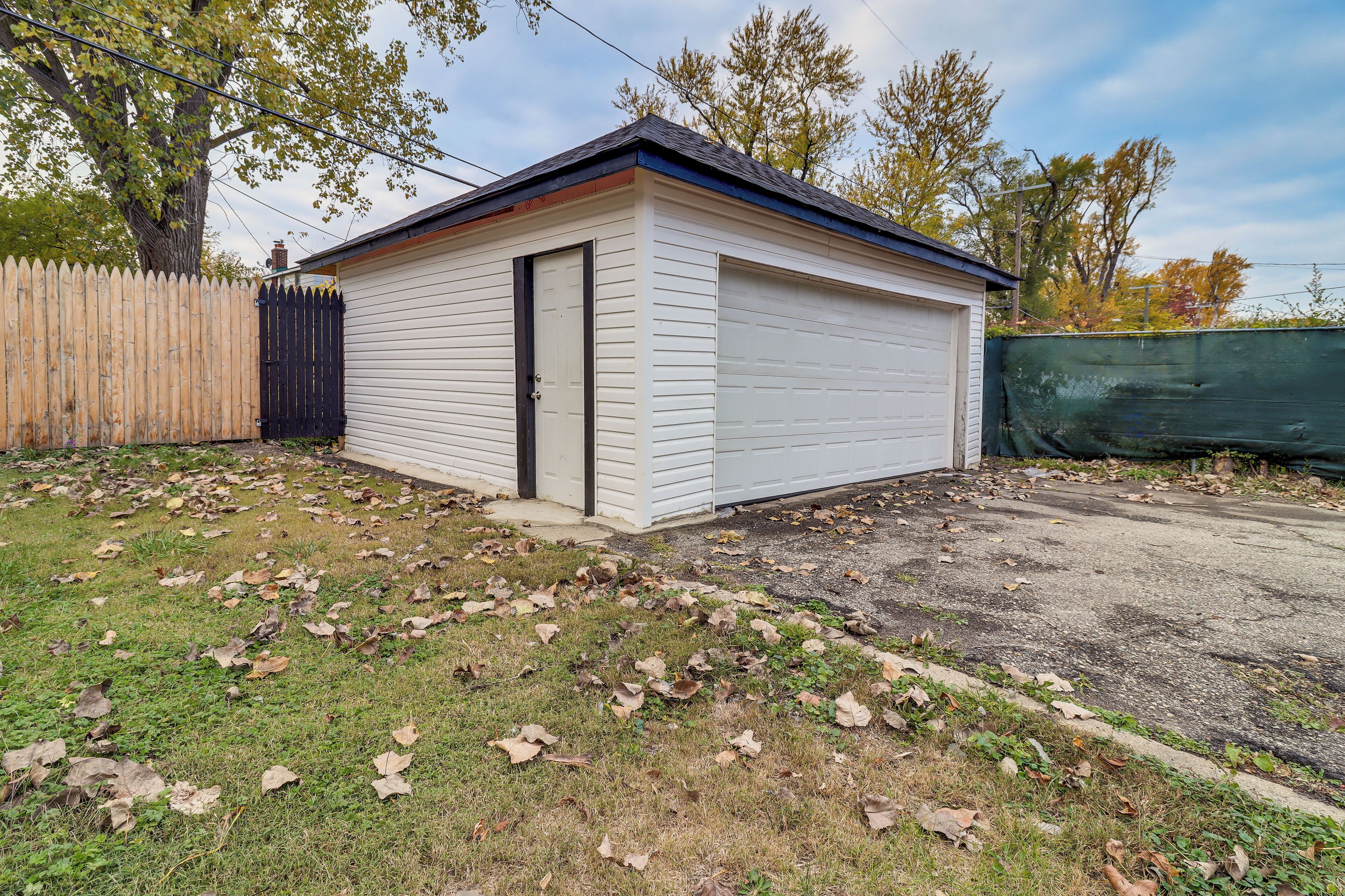 A small, single-story shed with a closed door, surrounded by a wooden fence and a grassy area with scattered leaves.