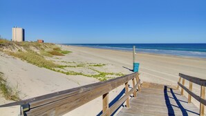 Une promenade en bois mène à une plage de sable, sous un ciel bleu dégagé, avec un bâtiment visible au loin.