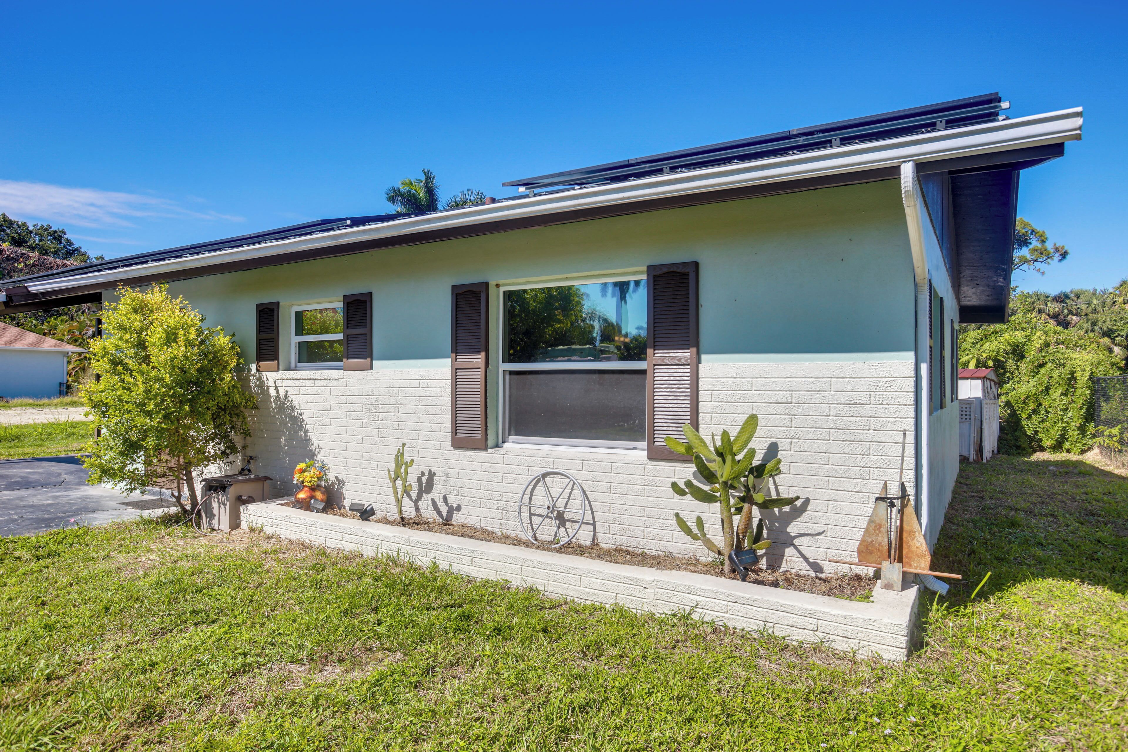 A modern house with a solar panel roof, a small garden with cacti and a wheelbarrow, and a clear blue sky.