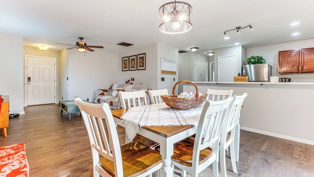 A dining area with a table set for four, surrounded by white chairs with wooden seats. A ceiling fan is visible above the table, and a basket with items is placed on the table. - Fenced-in Yard w/ Deck: Pet-friendly Lochbuie Home (Brighton)