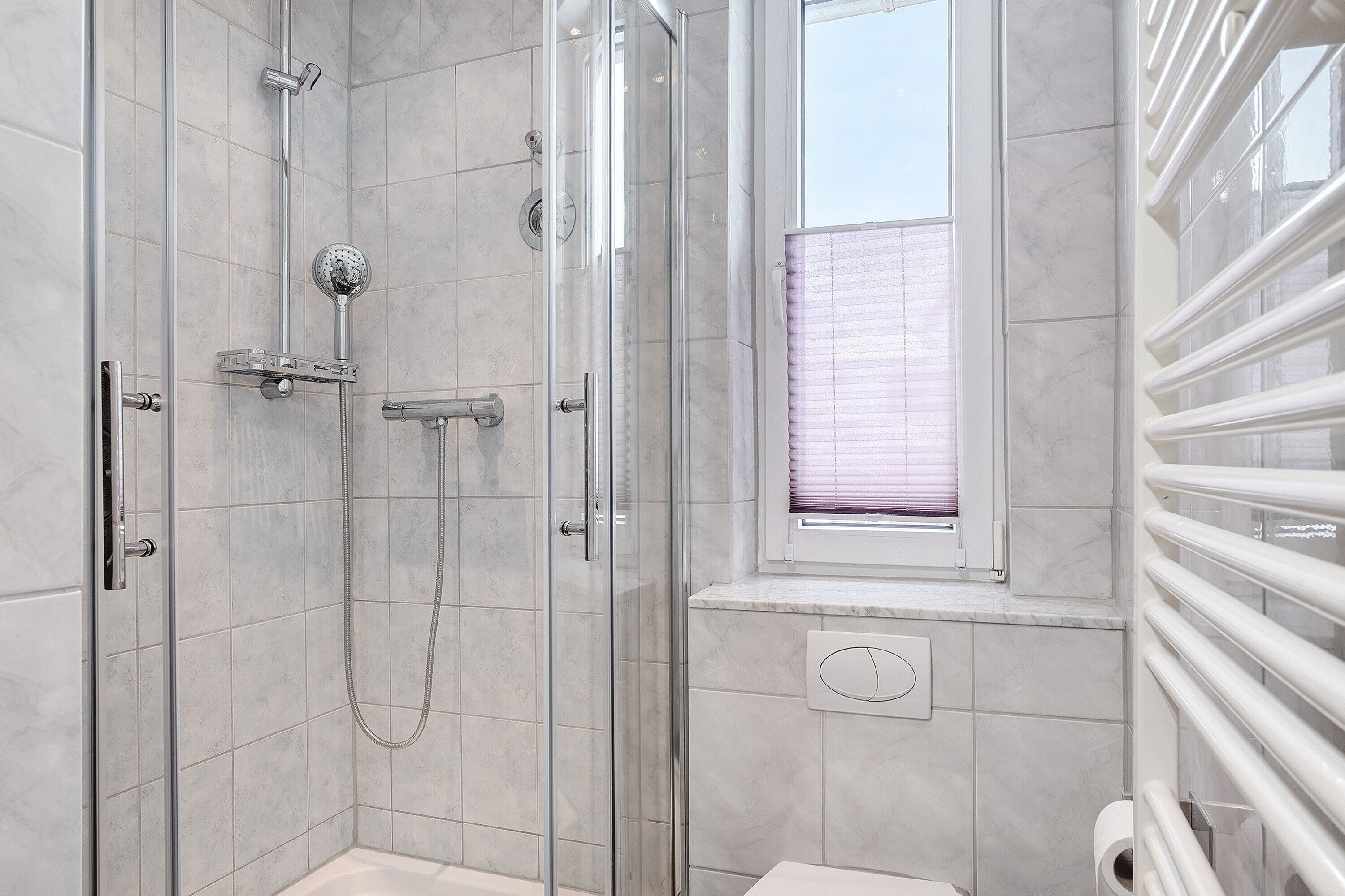 Un baño moderno con cabina de ducha de vidrio, una ventana con persianas y un radiador blanco.