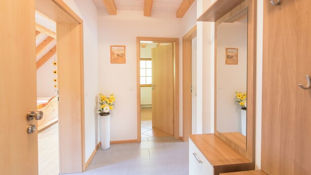 Um corredor com vigas de madeira, piso branco e um banco de madeira. Há dois arranjos florais sobre pedestais brancos.