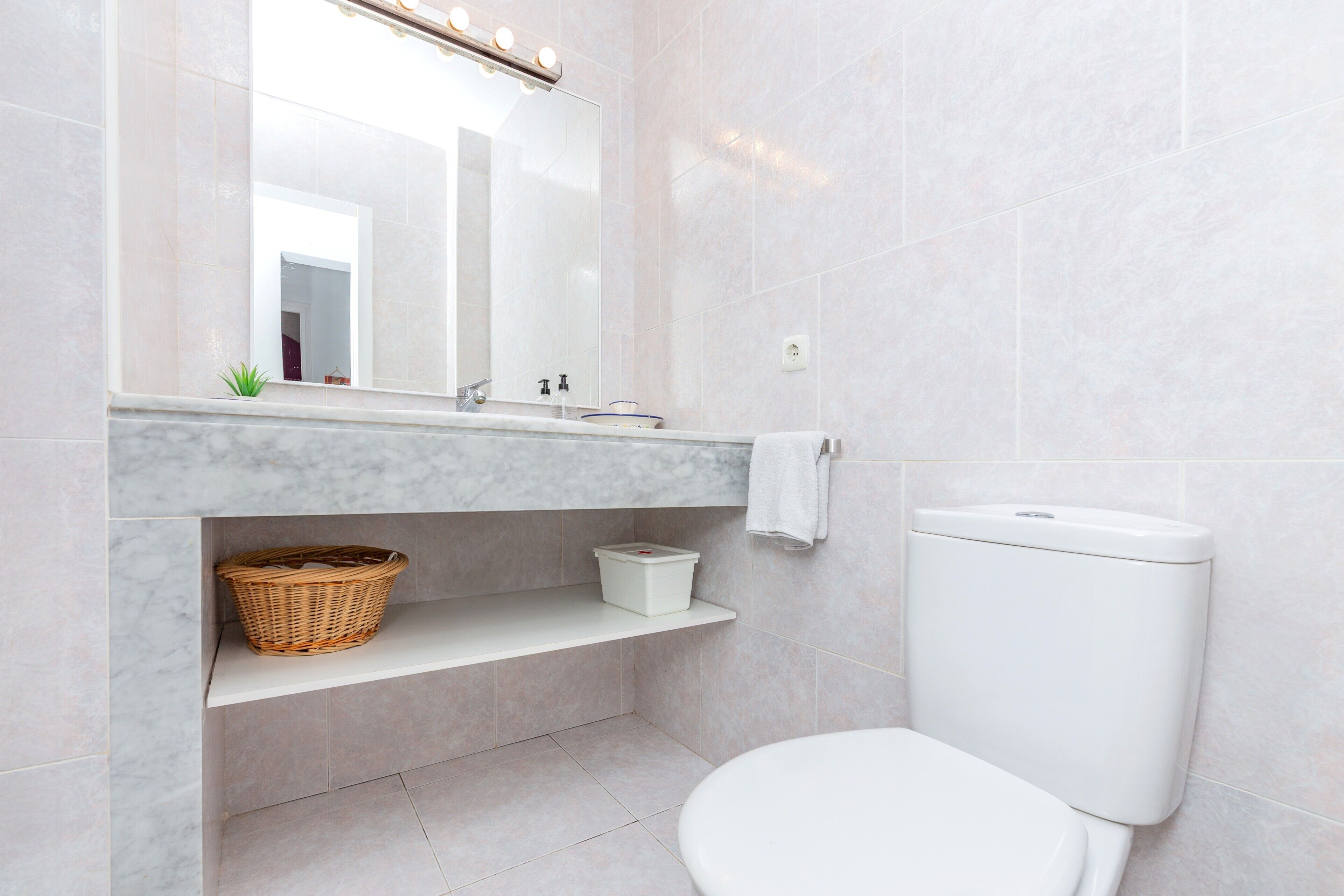 A bathroom with a white toilet, a mirror with lights, a shelf with a basket, and a marble countertop.