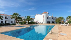 Una piscina con sillones de playa, un edificio blanco con tejado de tejas rojas y palmeras.