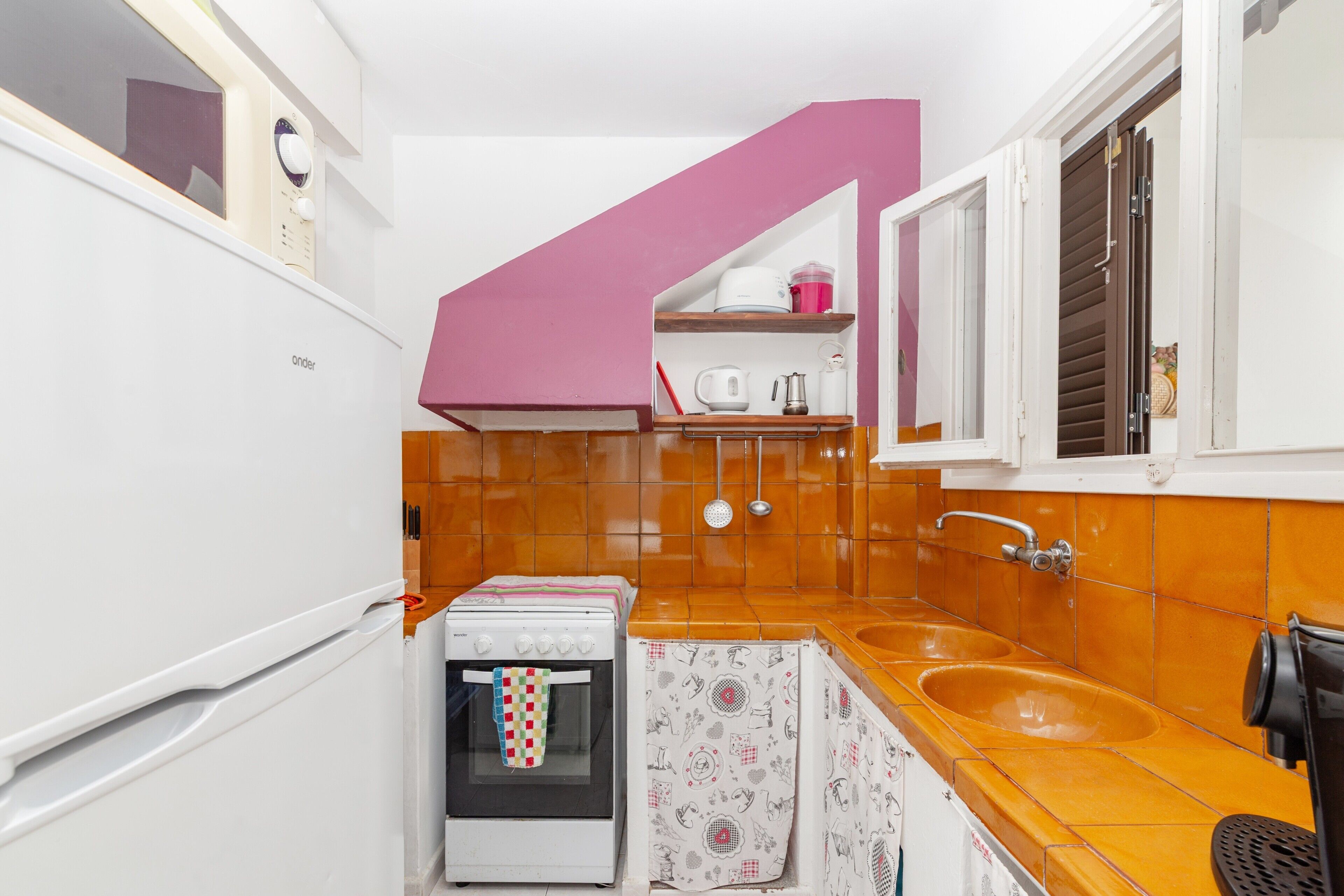 A compact kitchen with a white refrigerator, a small oven, a sink, and a window with blinds.