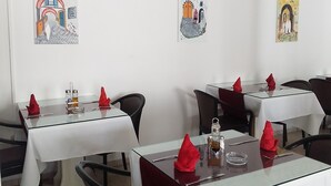 Un espace repas avec des tables dressées pour deux personnes, chacune agrémentée d’une nappe blanche, de serviettes rouges et de verres. Les murs sont décorés d’œuvres d’art encadrées représentant des scènes architecturales.