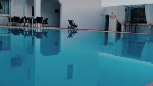 Une piscine à l’eau claire, entourée d’un bâtiment blanc doté d’un espace de repos couvert et de quelques chaises.