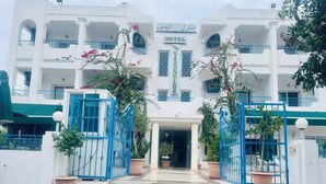 L’entrée d’un hôtel, avec une porte centrale encadrée de grilles bleues, des plantes en pot et une enseigne indiquant le nom de l’hôtel.
