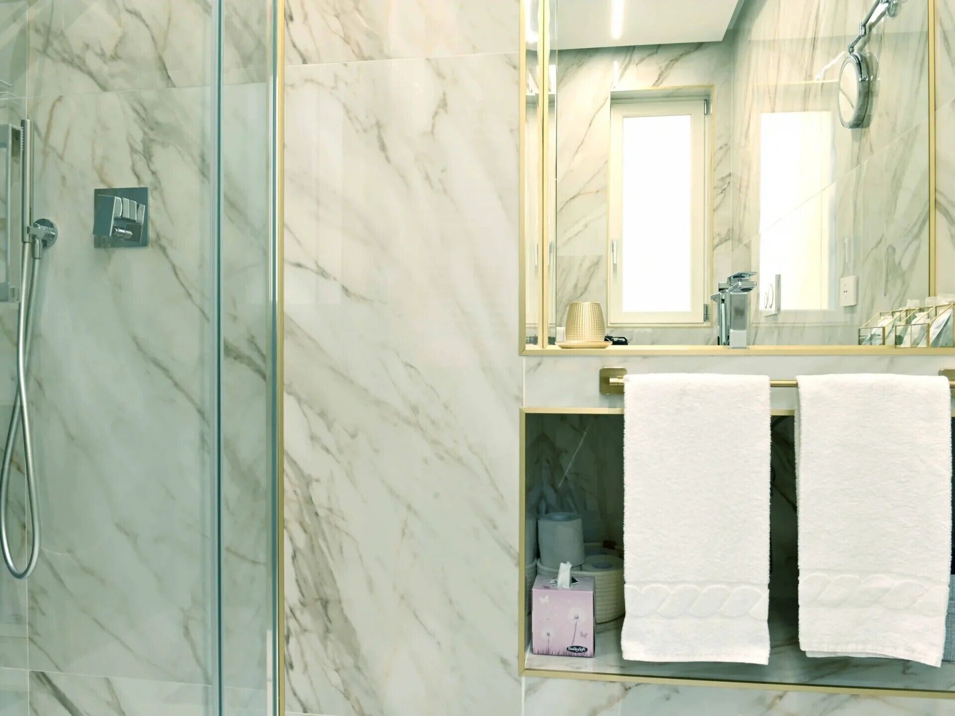 A bathroom with a glass shower enclosure, a marble wall, a mirror, and two white towels on a shelf.