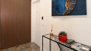 Un pasillo moderno con un revestimiento de listones de madera, una puerta blanca, una mesa de vidrio con una maceta roja y libros, y un gran cuadro abstracto en la pared.