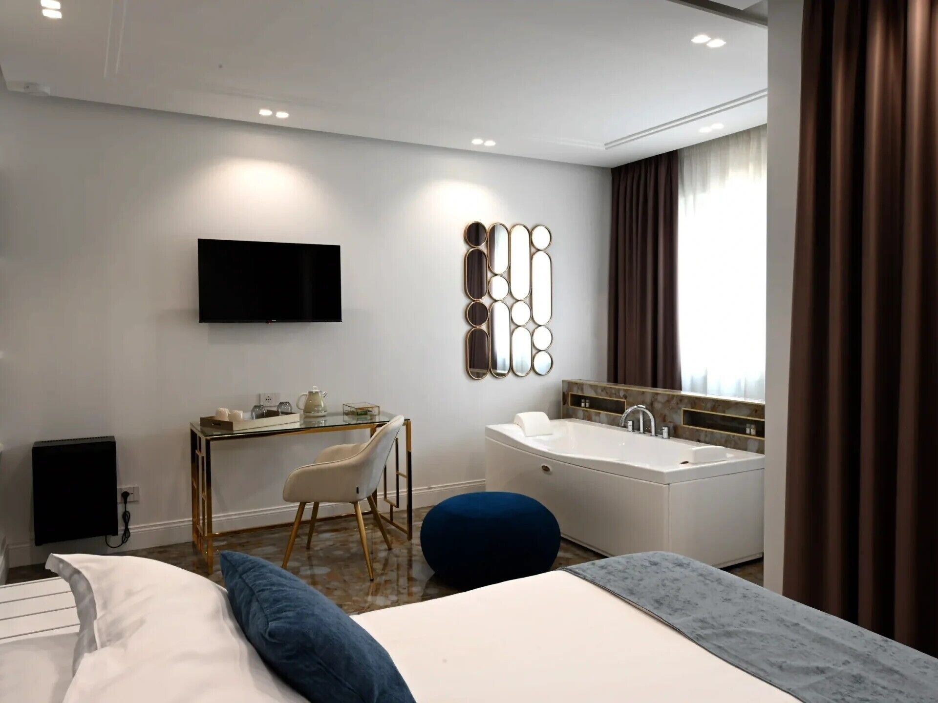 A modern hotel room with a bed, a desk, a flat-screen TV, a large mirror, a bathtub, and a window with curtains.