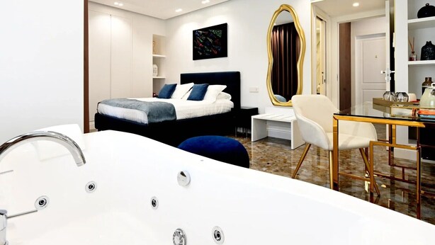 Un dormitorio moderno con una bañera blanca grande, una cama con ropa de cama azul, un escritorio con una silla y un espejo grande con marco dorado.