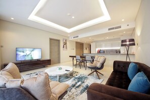 Ein modernes Wohnzimmer mit Flachbildfernseher, rundem Couchtisch und braunem Sofa mit blauen Kissen. Im Hintergrund ein Essbereich mit Tisch und Stühlen sowie eine Küche.