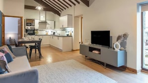 A modern living room with a grey sofa, a flat-screen TV on a grey console, a dining area with a table and chairs, and a kitchen in the background. - Swan Cottage (Gaerwen)