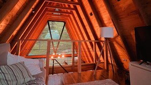 A cozy attic bedroom with a sloped ceiling, wooden beams, a bed, a desk, and a large window offering a view of the outdoors. - Exclusive A-Frame chalet with private pool. (Itajubá)