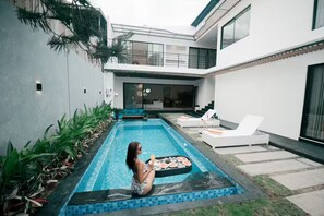 A modern outdoor pool area with a person relaxing on a lounge chair, surrounded by greenery and a dining setup. - Sale45% Villa 4BR @Seminyak near Double Six Beach (Seminyak)