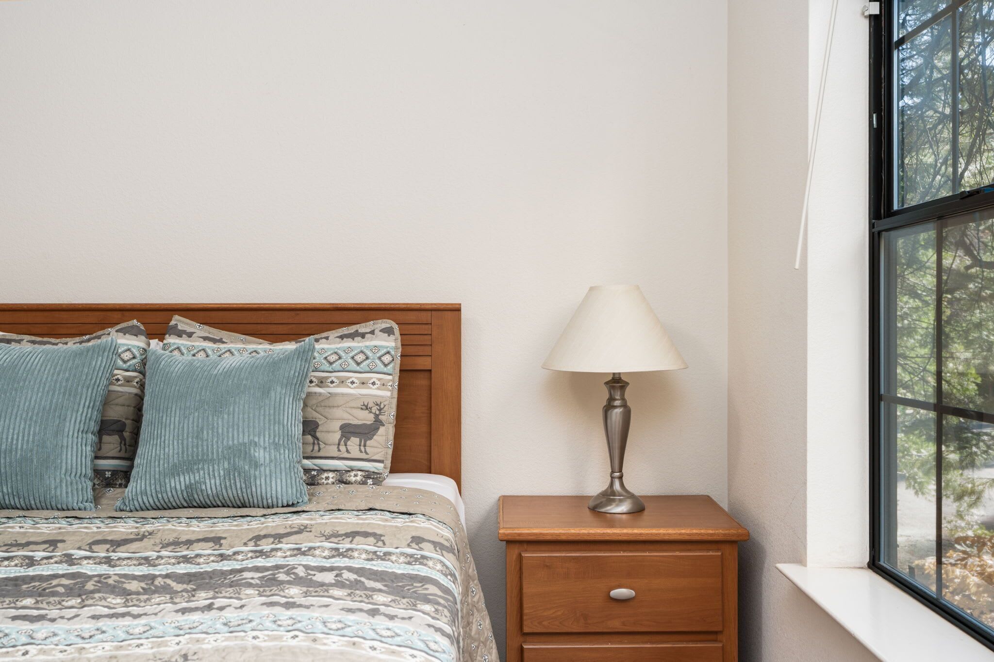 A bedroom with a bed, patterned bedding, a nightstand with a lamp, and a window with a view of trees.