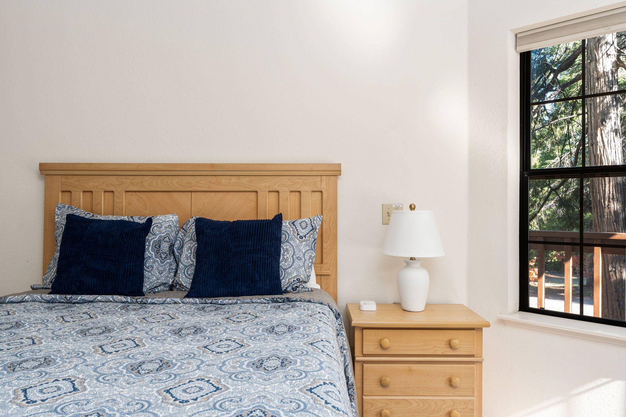A bedroom with a wooden headboard, a patterned bedspread, two pillows, a bedside table with a lamp, and a window with a view of trees.