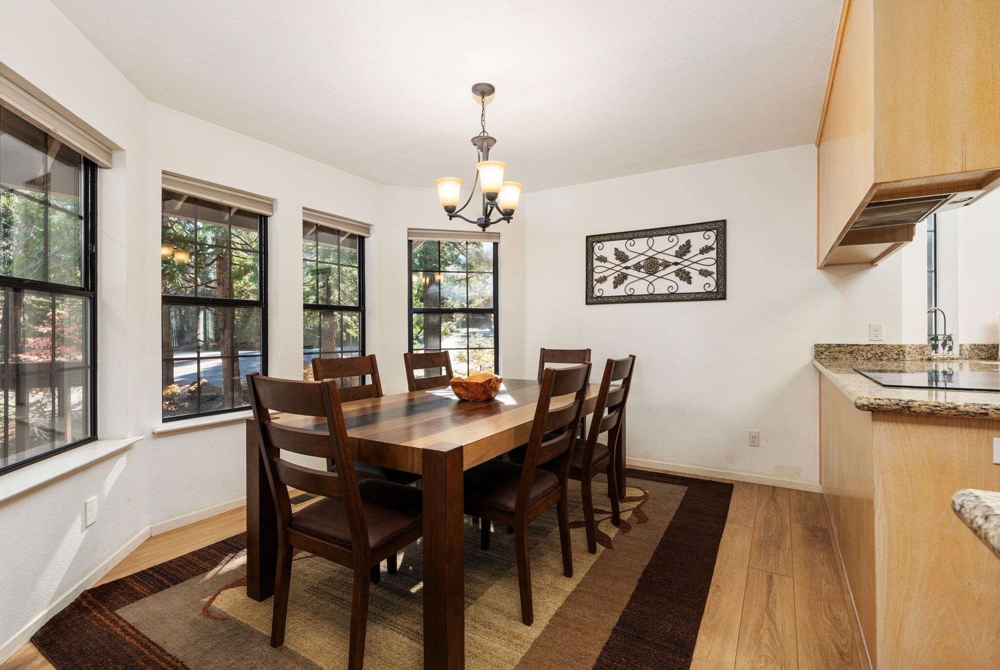 A dining area with a wooden table and chairs, a chandelier, and large windows with a view of trees.