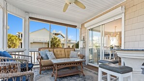 Un espacio exterior techado con mobiliario de mimbre, una mesa de cristal y un taburete blanco.
