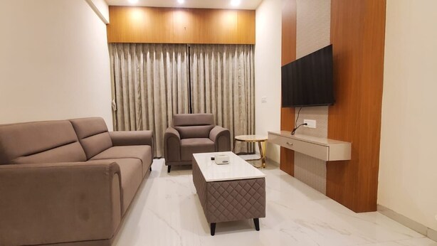 A modern living room with a sofa, armchair, and ottoman, a small round table, and a flat-screen TV mounted on the wall. - KRISHNA SAI SUITES (Rahata)