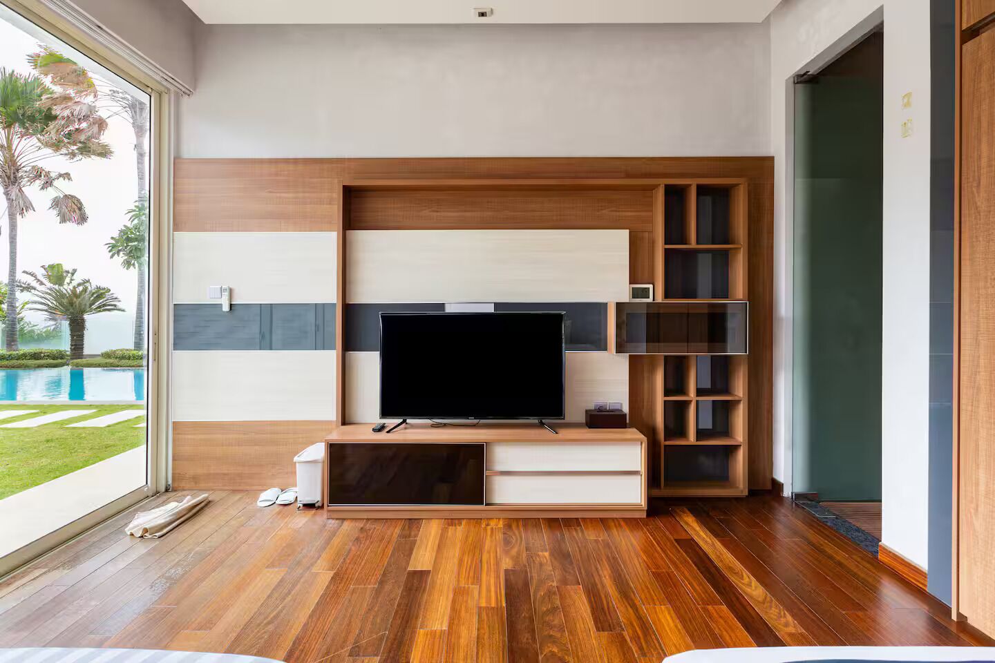 A modern living room with a flat-screen TV, wooden flooring, and a view of a swimming pool.