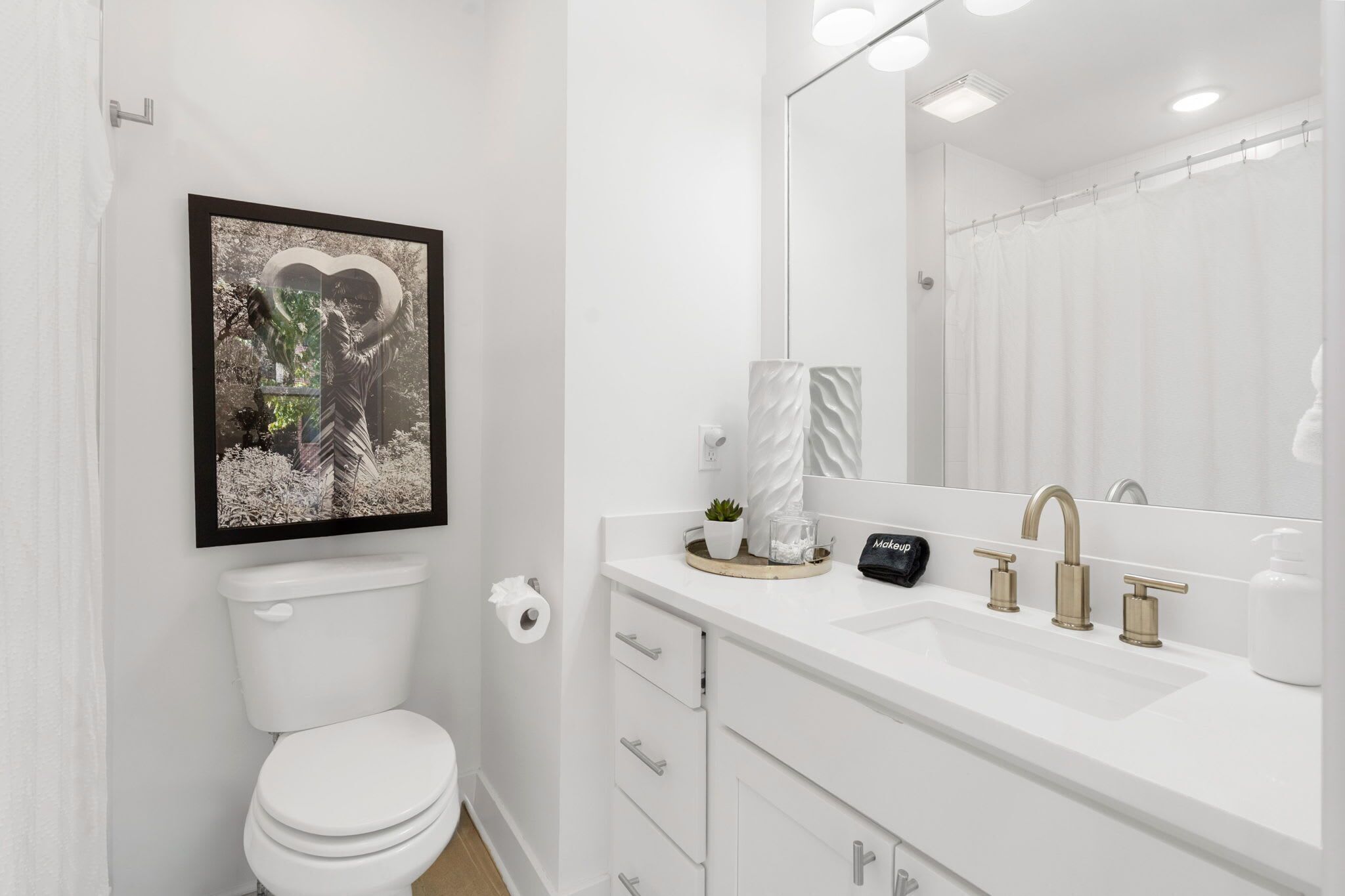 Un baño moderno con inodoro blanco, un lavamanos con un espejo grande y un cuadro en la pared.