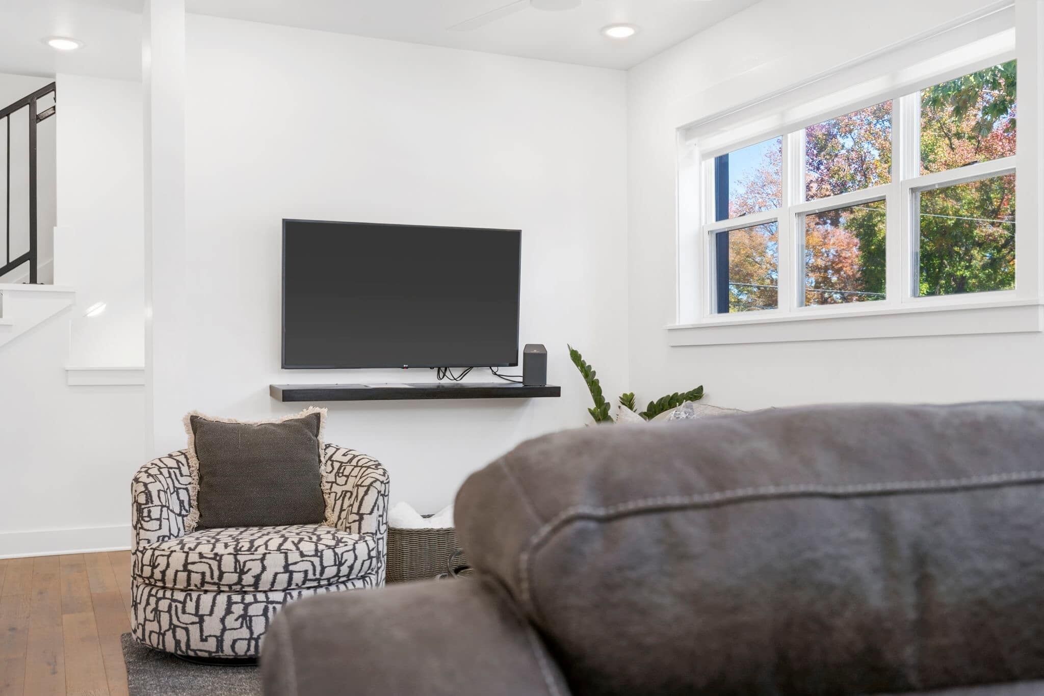 A living room with a modern design, featuring a large flat-screen TV mounted on the wall, a patterned armchair, and a window with a view of trees.