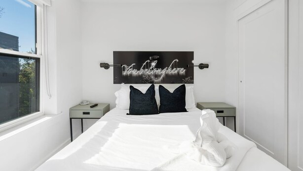 A modern bedroom with a large bed, a headboard with a motivational quote, two bedside tables, and a window providing a view of the outside. - G St Loft, Downtown Bentonville (Bentonville)