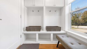 A clean, white locker room with two baskets on a bench, a bench, and a window with a view of trees and a fence. - G St Loft, Downtown Bentonville (Bentonville)