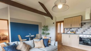 Uma sala de estar moderna com um sofá cinzento, almofadas azuis e uma mesa de centro redonda. É visível uma área de refeições com uma mesa pequena e cadeiras. A cozinha possui armários de madeira e um fogão.