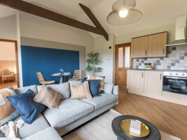 Una sala moderna con un sofá gris, cojines azules y una mesa de centro redonda. Se observa un comedor con una mesa pequeña y sillas. La cocina tiene armarios de madera y una estufa.