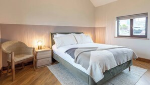 Um quarto com cama, mesa de cabeceira, luminária, cadeira e janela.