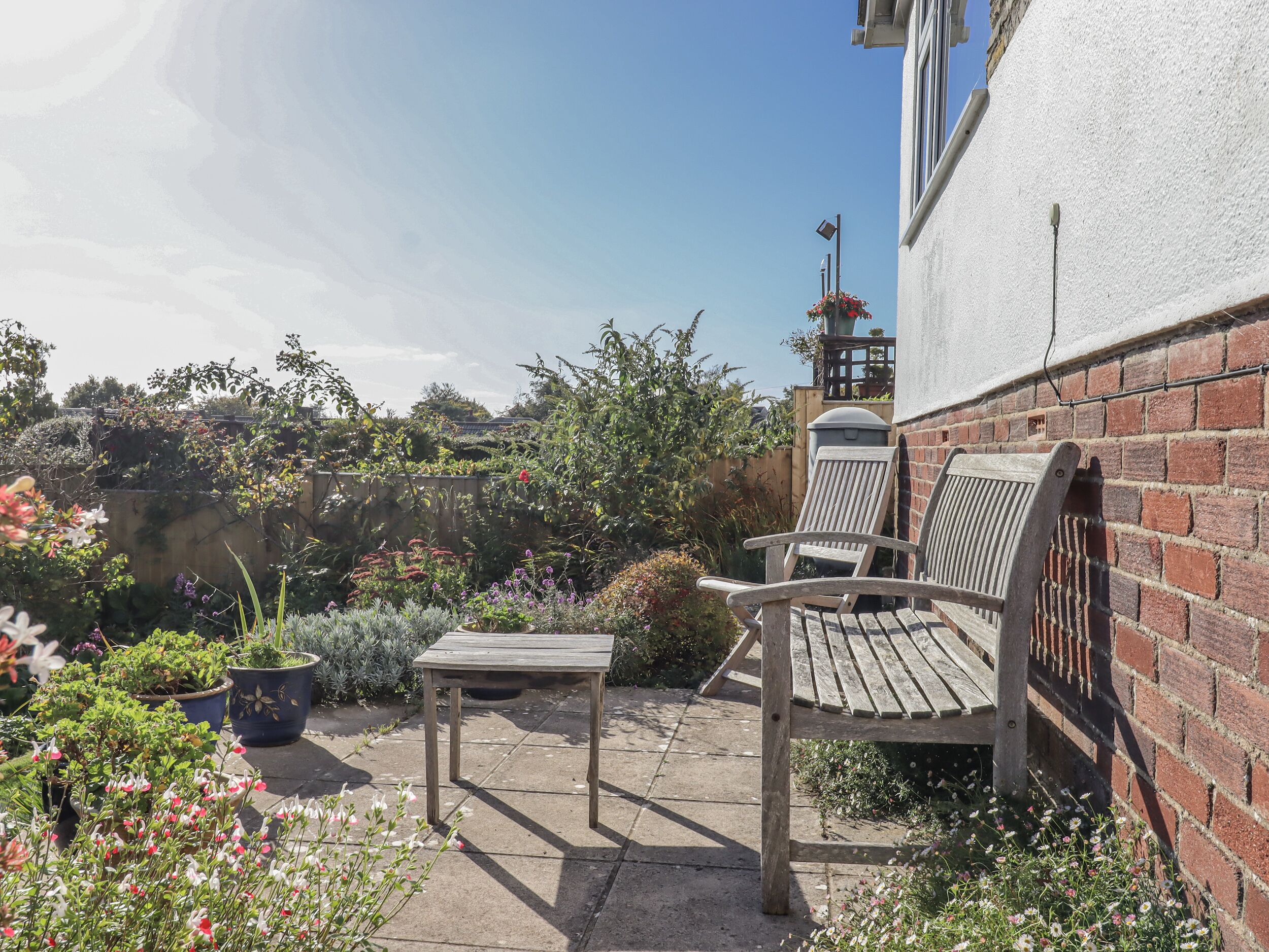 A patio with a wooden table and two wooden chairs, surrounded by potted plants and flowers.