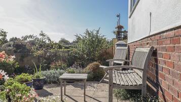 A patio with a wooden table and two wooden chairs, surrounded by potted plants and flowers.