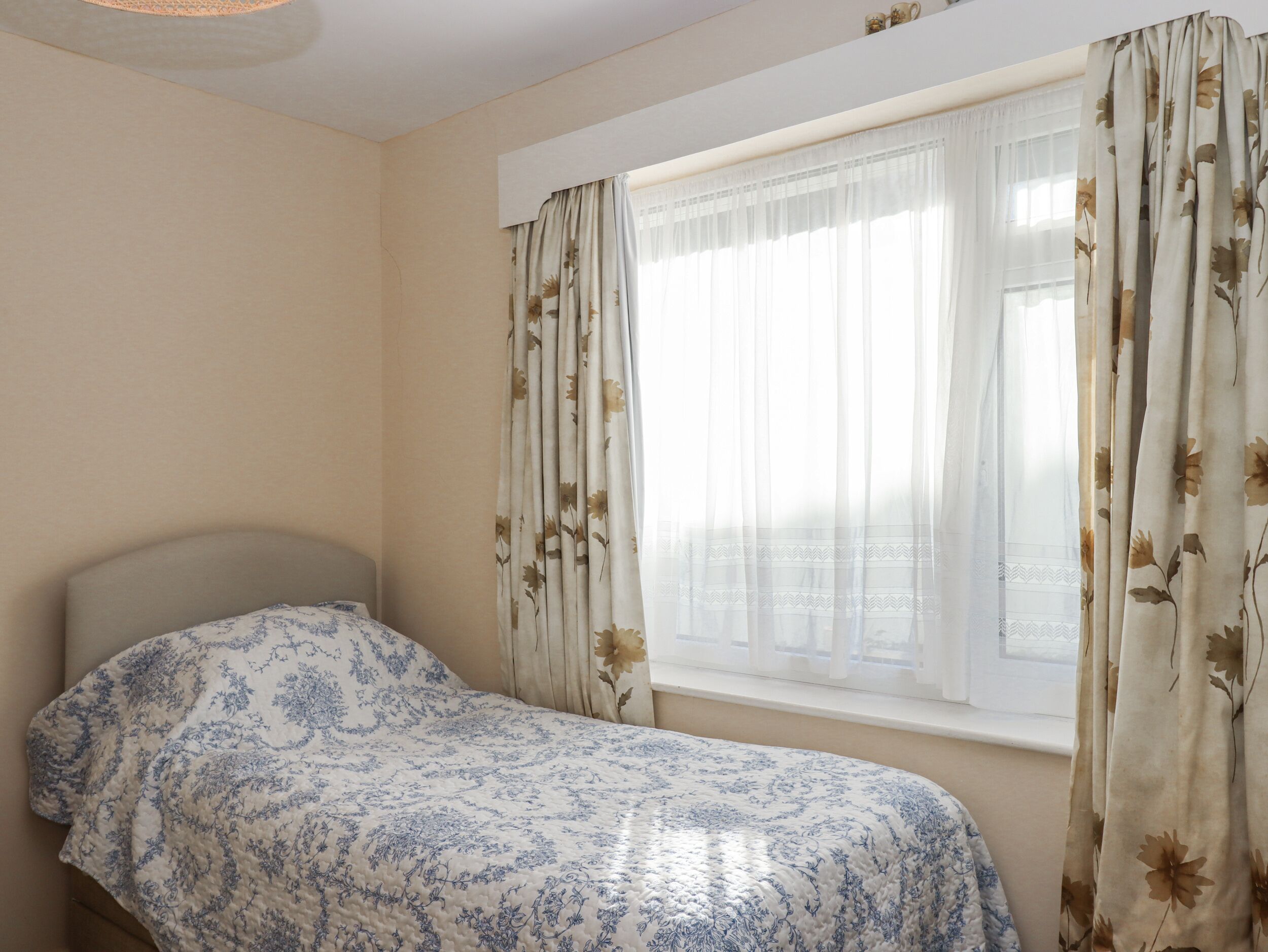Une chambre avec un lit simple, une fenêtre ornée de rideaux à motifs floraux et un mur uni.