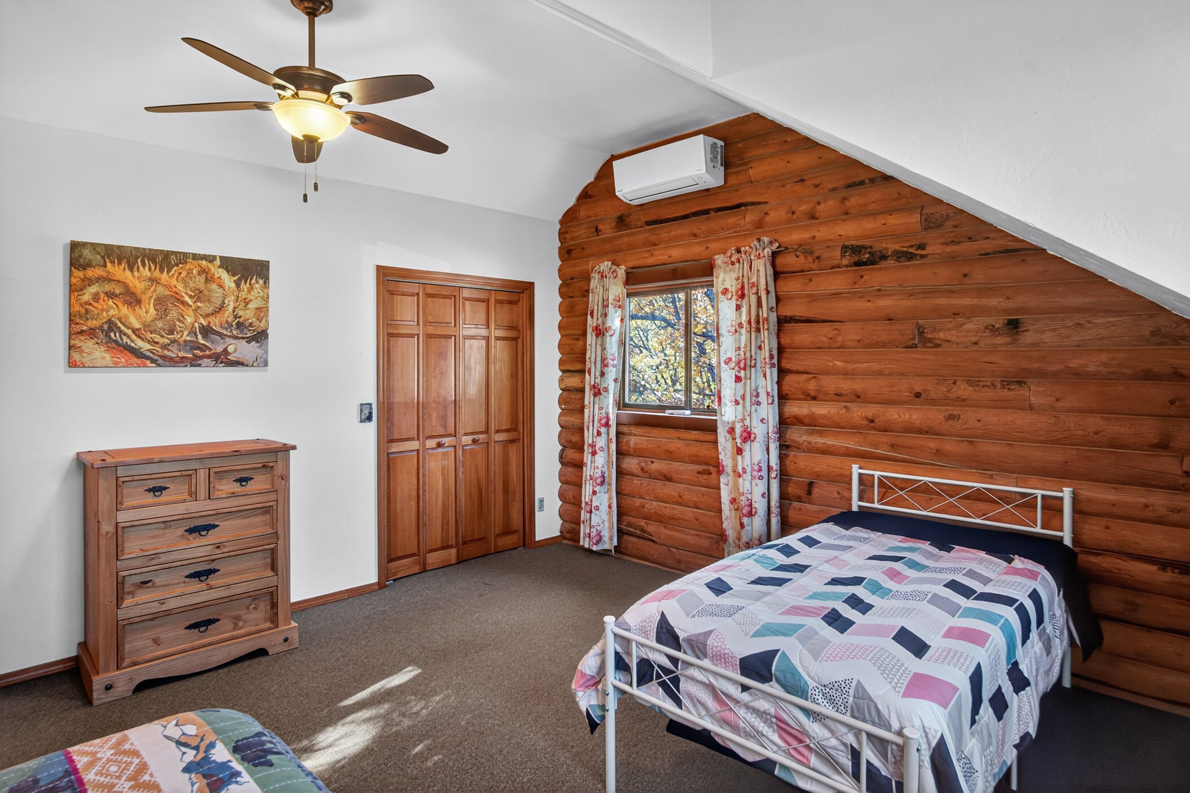 A bedroom with a sloped ceiling, wooden walls, a ceiling fan, a bed with a quilt, a wooden dresser, a door, and a window with curtains.