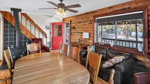 A cozy cabin interior with a dining table, chairs, a sofa, a fireplace, and a large window offering a view of the outdoors. - Minnie Mine Cabin Retreat 4 Bedroom Cabin (Prescott)