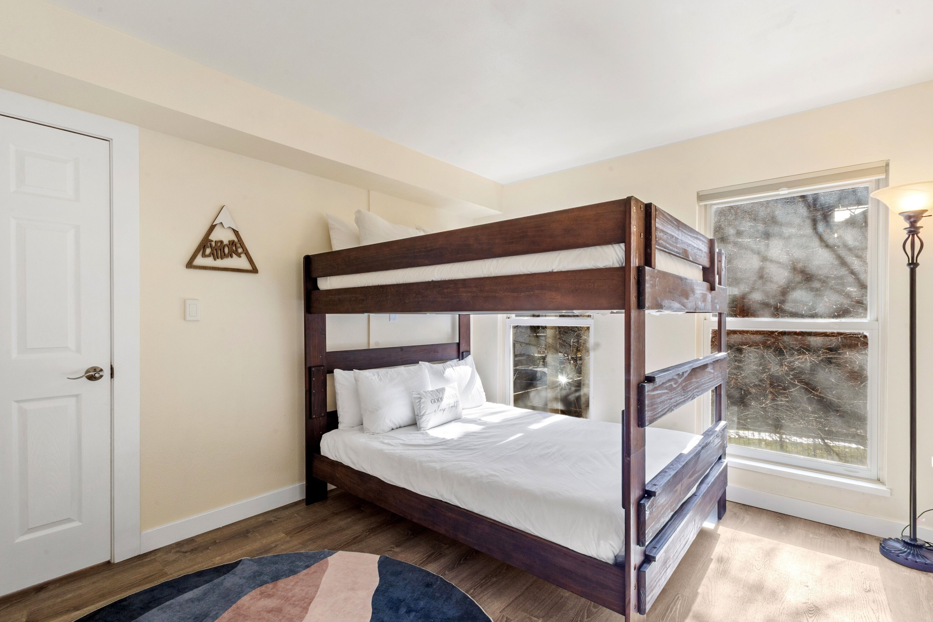 A bunk bed with white bedding, a wooden floor, a white door, a wall-mounted decoration, and a window with a view of trees.