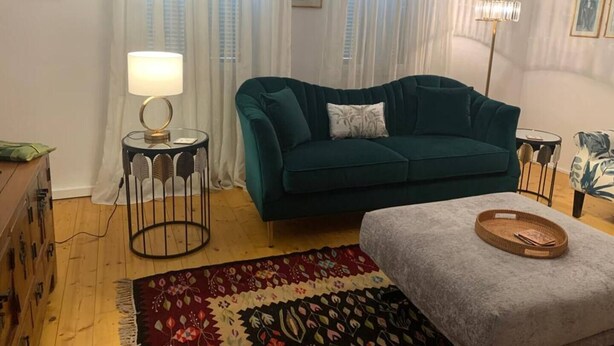 Una sala de estar con un sofá azul aguamarina, un tapete estampado, una pequeña mesa redonda con una lámpara y una mesita auxiliar con lámpara.