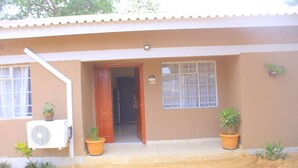 A building with a brown exterior, a front door, two potted plants, and an air conditioning unit. - Blue Ridge Escape Guest House (Kasane)