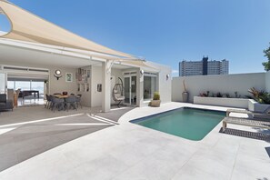 A modern outdoor pool area with a covered seating area, a dining table, and a view of the city skyline. - Luxury Family Villa - Ocean & Mountain Views (Bloubergstrand,)