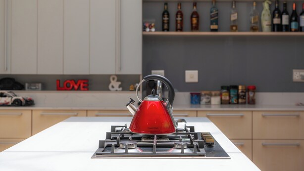 Een rode waterkoker op een fornuis in een keuken, met flessen op de achtergrond.