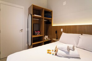 Habitación de hotel con cama, ropa de cama blanca, armario de madera, mesita de noche y espejo adosado a la pared.