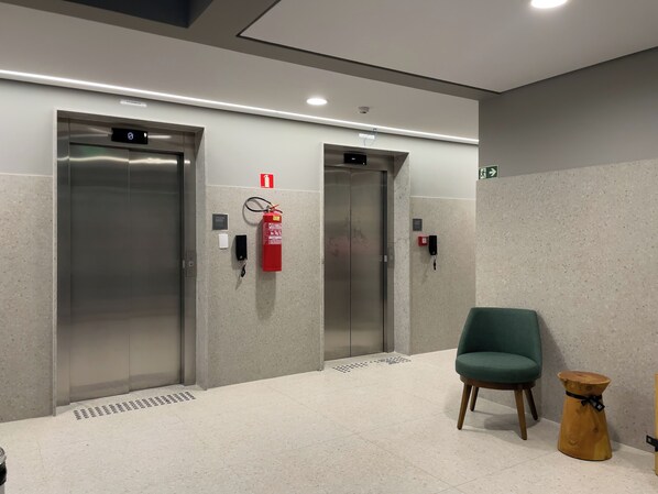 An elevator area with two elevators, a fire extinguisher, a phone, and a green chair. - Virgílio Pinheiros (São Paulo)