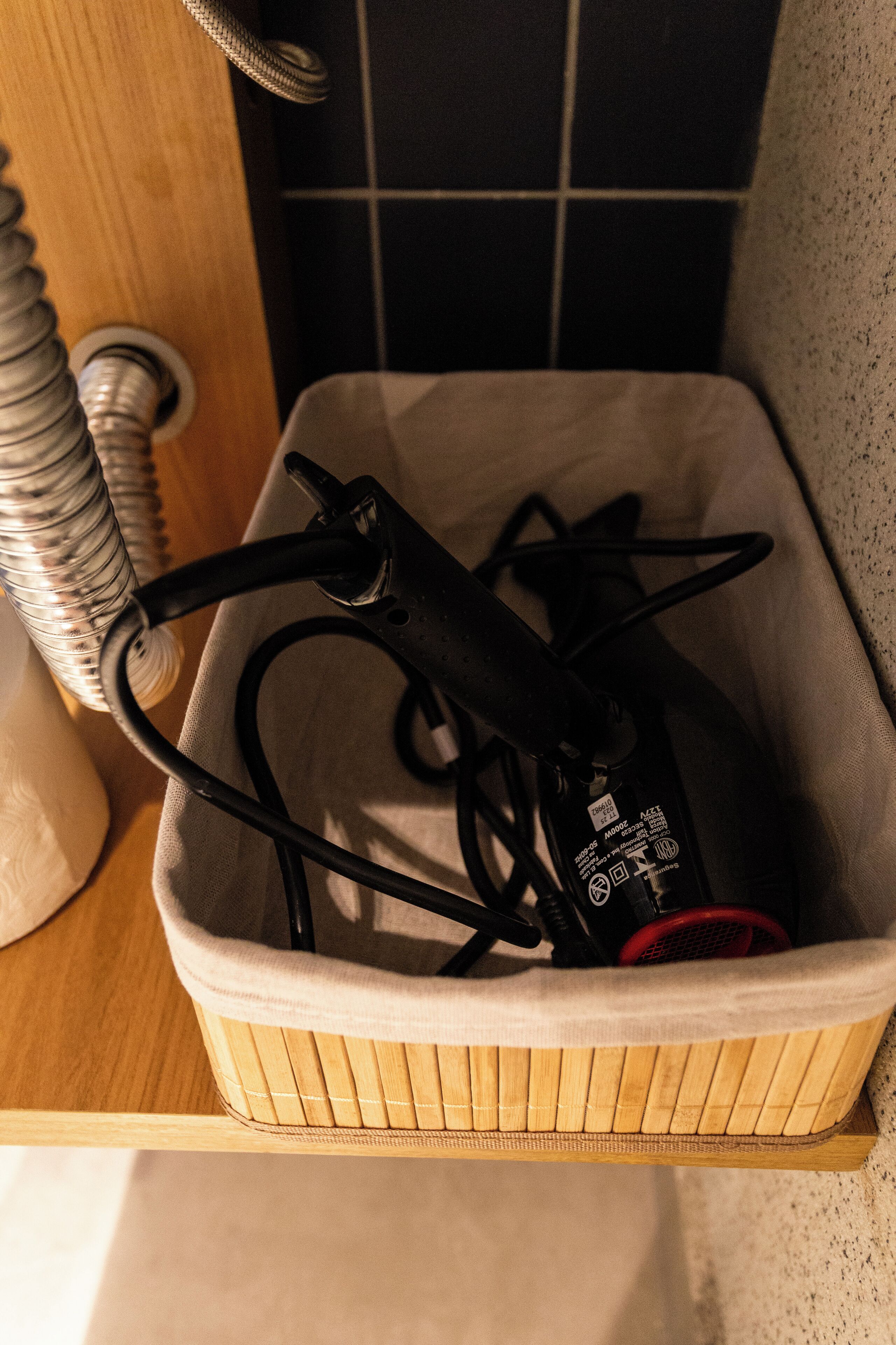 A wooden basket with a white cushion, containing a black power strip and a red object, possibly a power adapter, on a wooden surface with a tiled wall in the background.