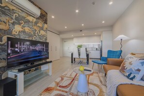 A modern living room with a flat-screen TV, a sofa, a coffee table, and a dining area in the background. - Stylish CBD Stay | 2BR | Private Deck (Christchurch)