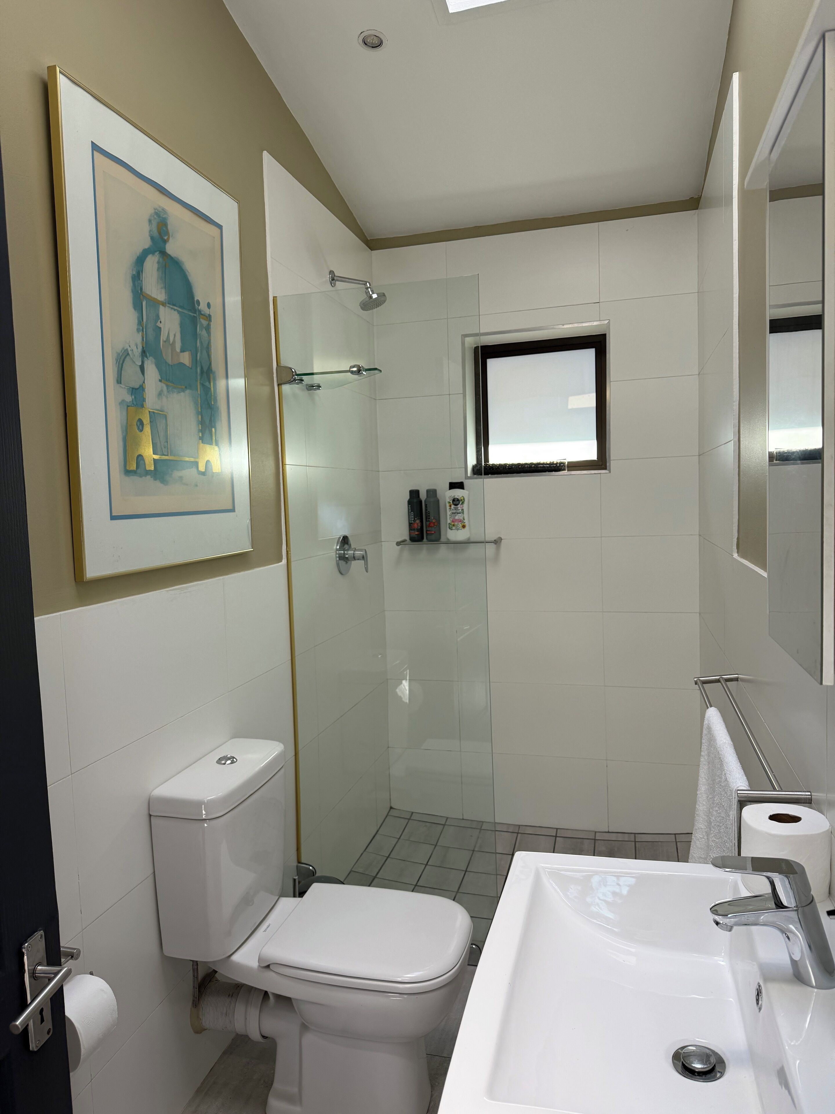 A modern bathroom with a toilet, sink, shower, and a framed artwork on the wall.