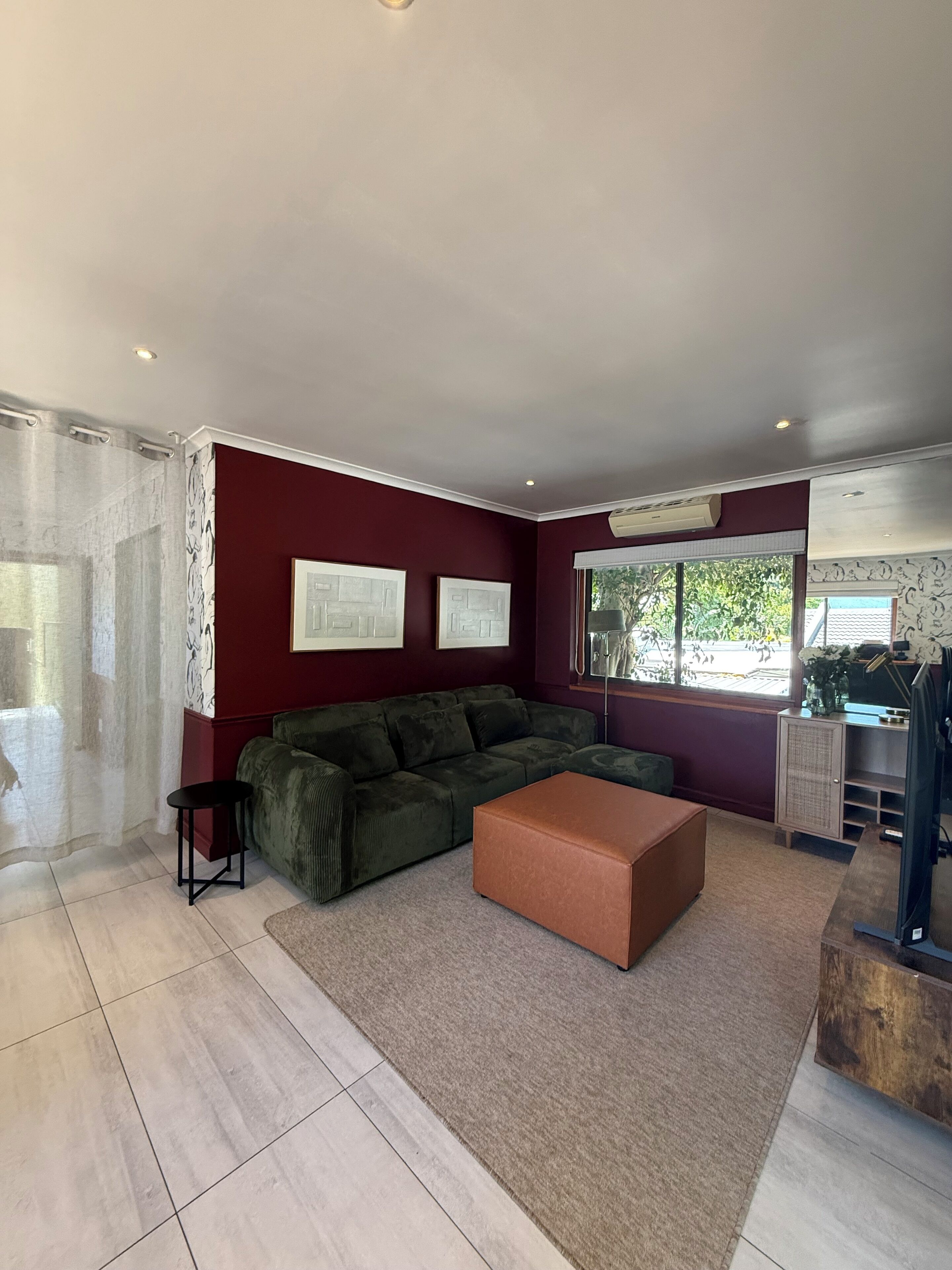 Una sala de estar moderna con una pared de acento rojo oscuro, un sofá verde, un reposapiés marrón y un ventanal con vistas a la vegetación.