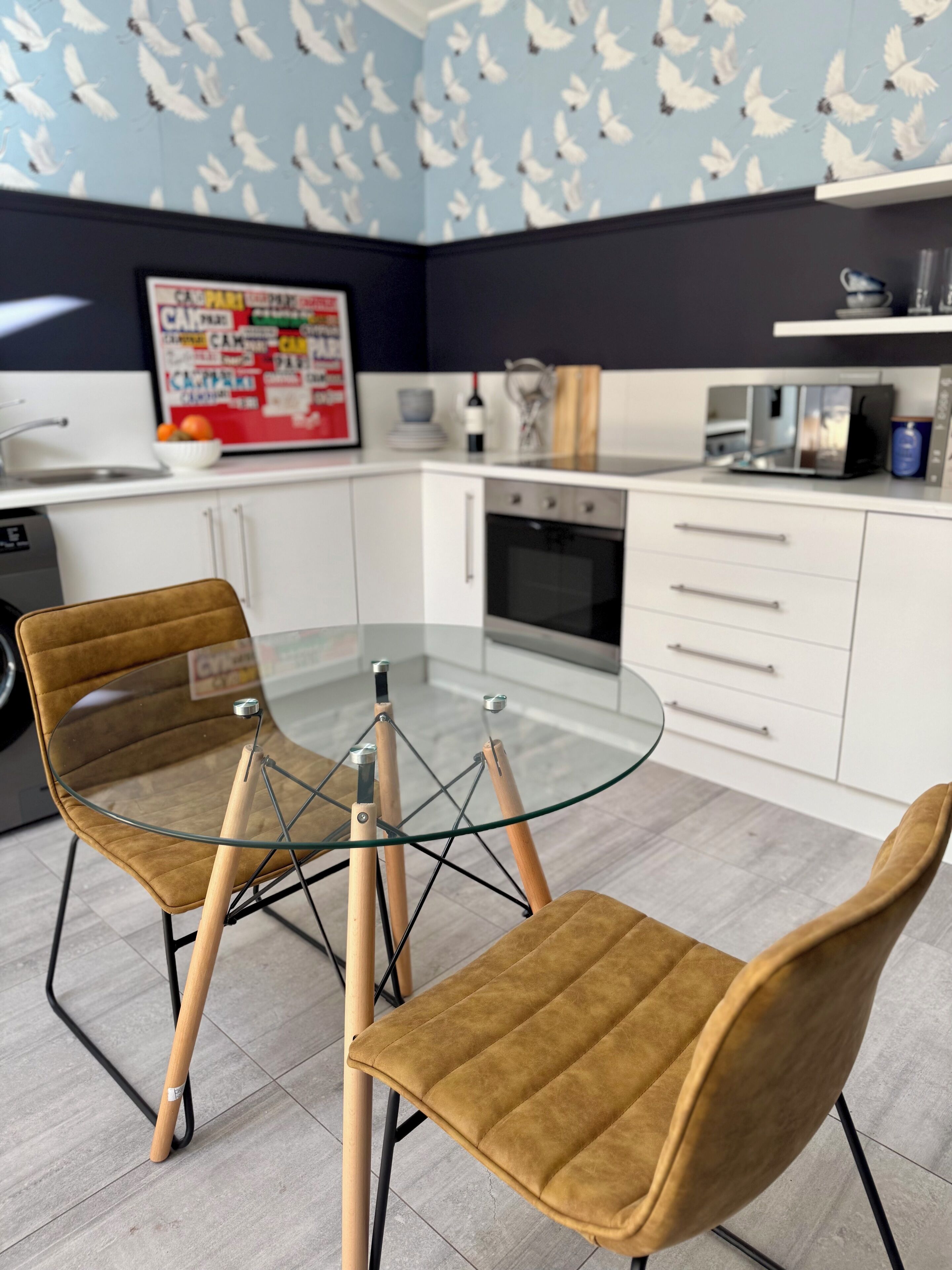 A modern kitchen with white cabinets, a glass-top table, and two brown chairs.